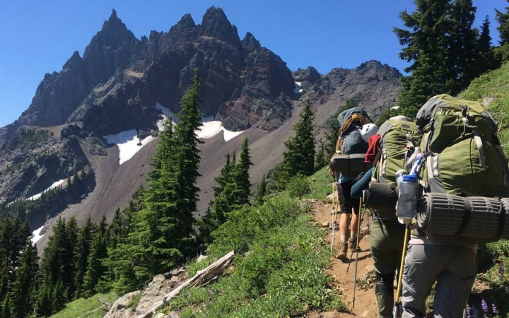 A group of students wearing backpacks hike along a trail toward a rocky, mountainous landscape